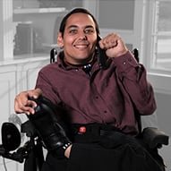 A young adult male with limited mobility wearing a maroon shirt smiling.